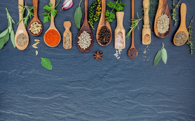 Photo various of spices and herbs in wooden spoons on the black stone.