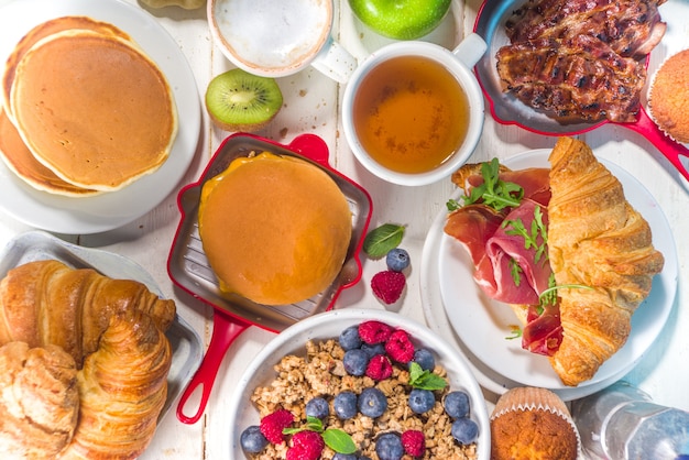 Various traditional breakfast food - fried eggs with bacon,\
muesli, oats, waffles, pancakes, burger, croissants, fruit berry,\
coffee, tea and orange juice, white table background copy space top\
view