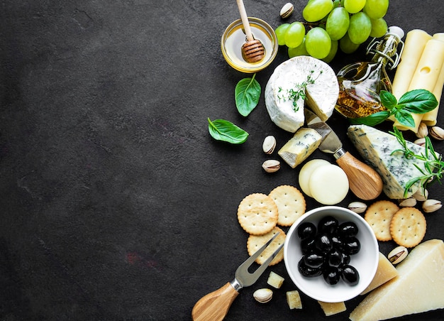 Various types of cheese, grapes, honey and snacks on a black concrete background