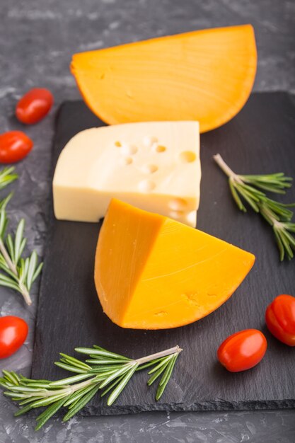 Various types of cheese with rosemary and tomatoes on black slate board on a black concrete surface