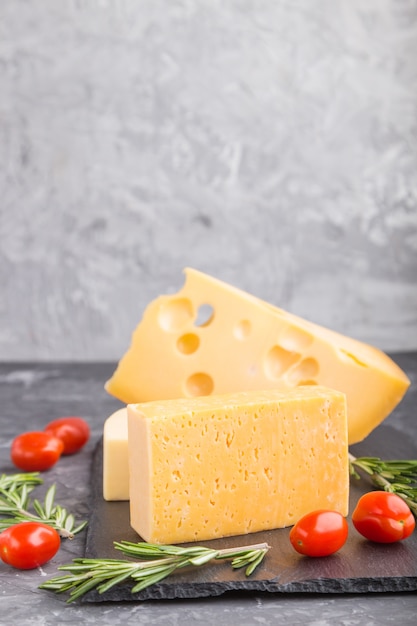 Various types of cheese with rosemary and tomatoes on black slate board on a black concrete table. Side view, close up, copy space.