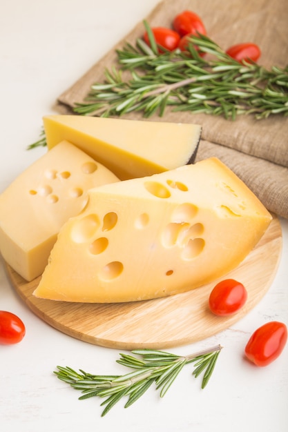 Various types of cheese with rosemary and tomatoes on wooden board on a white wooden surface. Side view, close up.