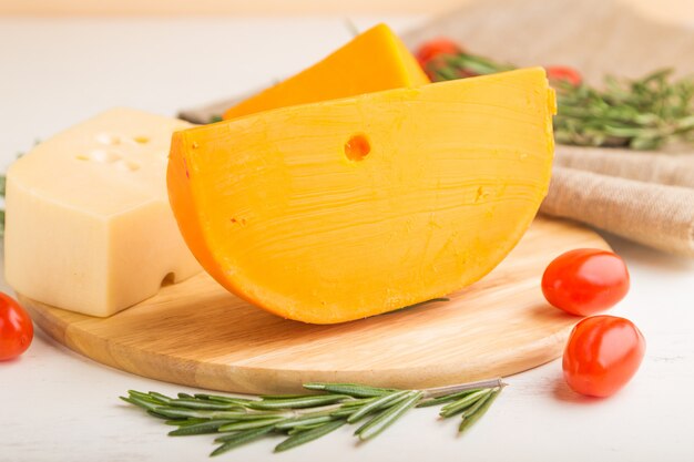 Various types of cheese with rosemary and tomatoes on wooden board on a white wooden surface. Side view, close up.