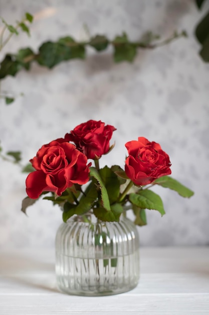 A vase of red roses is on a table with a wall behind it.