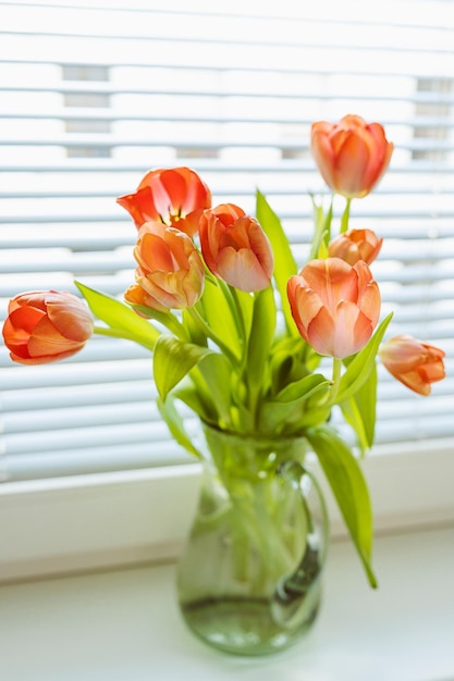 A vase of tulips sits on a window sill with a white blinds.