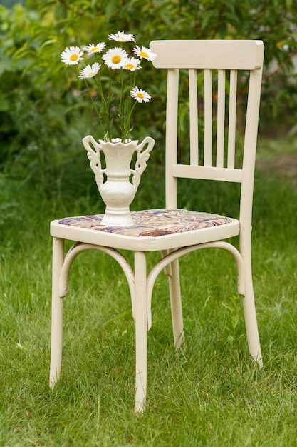 Photo vase with chamomile flowers on a white rustic styled chair in the spring garden with green grass