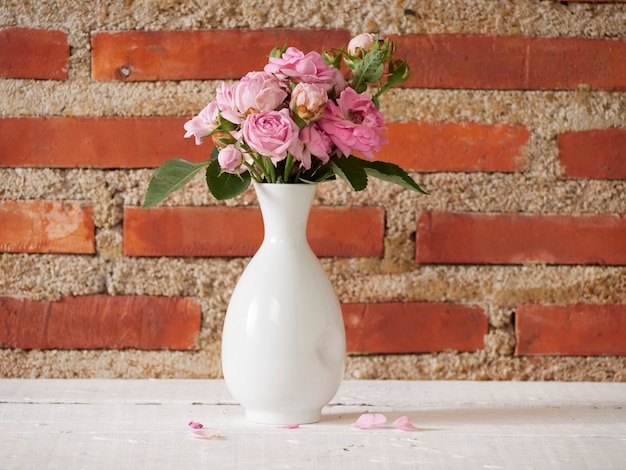 Vase with pink roses on white table near brick wall