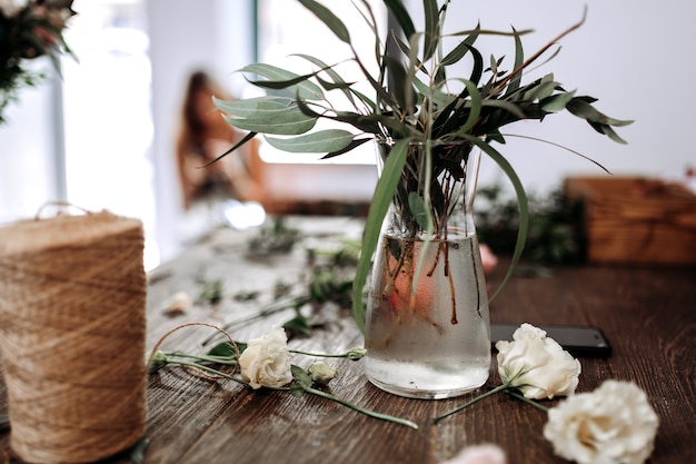 A vase with water and branches with green leaves and bobbin of twine on the table of florist