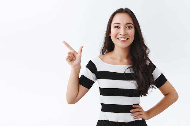 Vastberaden knappe aziatische vrouw in gestreept t-shirt beveelt product aan houd de hand op de heup zelfverzekerd wijzend naar de linkerbovenhoek en glimlachend levendig advies gevend kies iets in de winkel