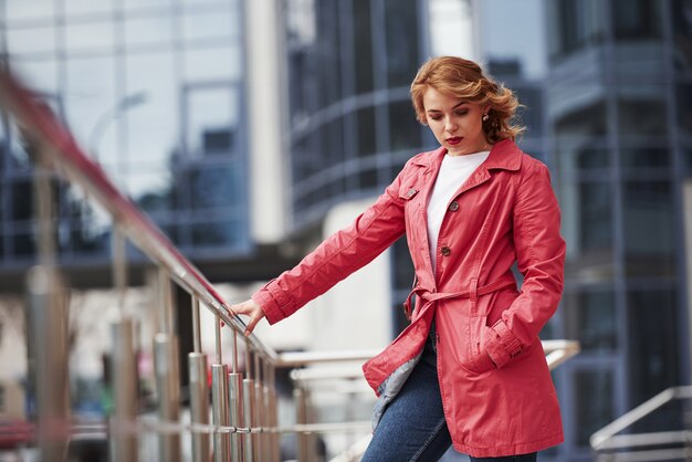 Vasthouden aan balustrades. Volwassen mooie vrouw in warme rode jas wandelt in het weekend door de stad