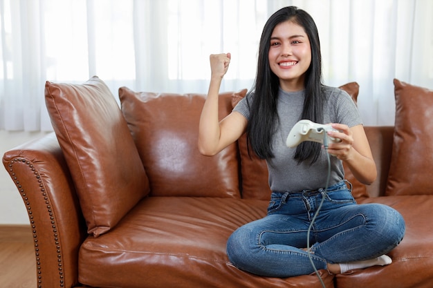 Фото Концепция игровой консоли vdo. активная азиатская женщина сидит на диване, держит джойстик и играет в увлекательную игру. симпатичная девушка выглядела взволнованной с игровой консолью