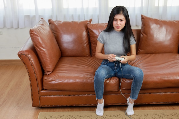 Vdo game console station concept. active asian woman sitting on\
sofa, holding joystick and playing exciting game. cute girl looked\
excited with game controller console