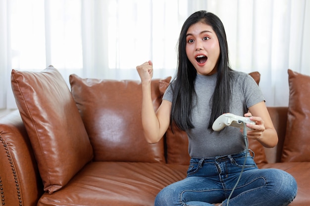 VDO game console station concept. active asian woman sitting on sofa, holding joystick and playing exciting game. Cute girl looked excited with game controller console