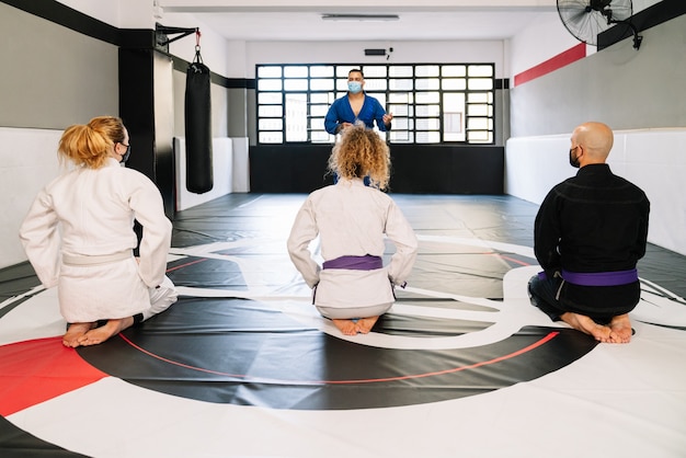 Foto vechtsportleraar die de beste technieken uitlegt terwijl studenten geknield op de mat worden gehoord terwijl ze gezichtsmaskers dragen vanwege het covid19 coronavirus