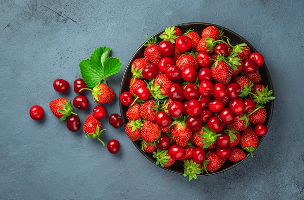 Veel kersen en aardbeien in een bord op een grijsblauwe achtergrond. Gezonde bessen en fruit.