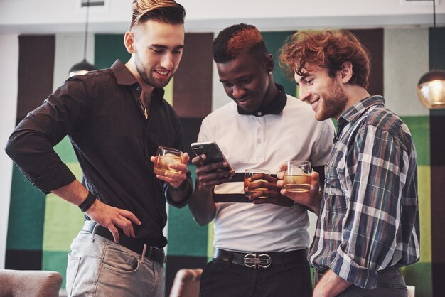 Veelbelovende jonge multi-etnische mensen vieren en drinken whisky toast, kijken gadget