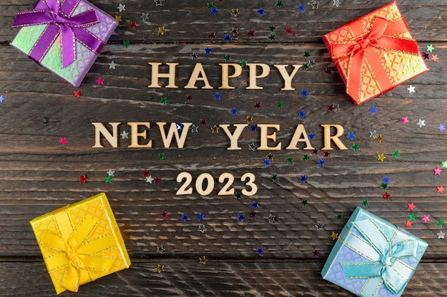 Veelkleurige geschenkdozen met gebonden strikken Nieuwjaar presenteert op houten tafel met verspreide confetti sterren Tekst Gelukkig Nieuwjaar 2023 Feestelijke wenskaart Plat lag