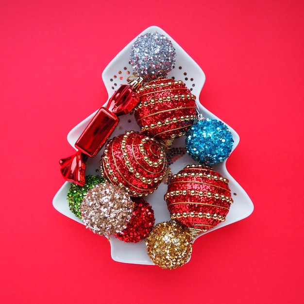 Veelkleurige kerstballen op een witte plaat in de vorm van een kerstboom Kerstboom Rode achtergrond of karmozijnrode kleur Mooi gestreept kerstdecor en glitters Plat lag Stilleven