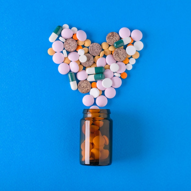 Veelkleurige tabletten worden uit een glazen vat in de vorm van een hart op een blauwe achtergrond gegoten. Het uitzicht vanaf de top. Het concept van behandeling en preventie van ziekten. Plat liggen.