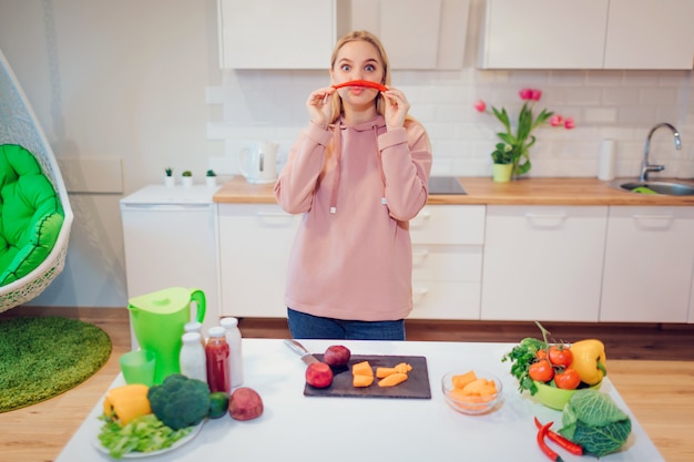 Vegan beautiful blonde woman has fun with chili pepper while cooking raw vegetables in the kitchen.