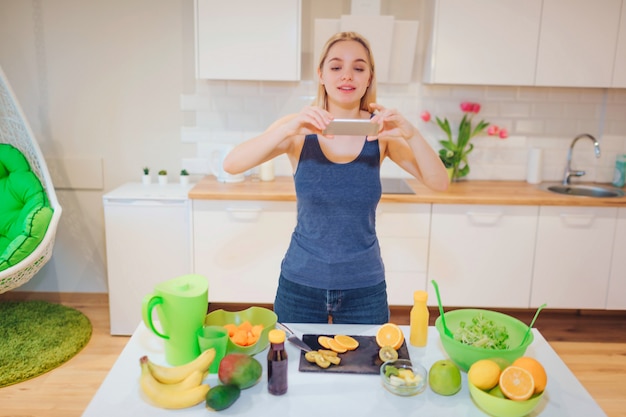 Vegan beautiful blonde woman with smartphone photographing organic orange in the kitchen