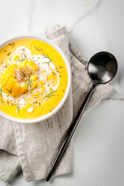 Vegan cuisine. Traditional autumn corn soup. Served with corn cobs, greens, cream. On a white marble table. Copy space top view