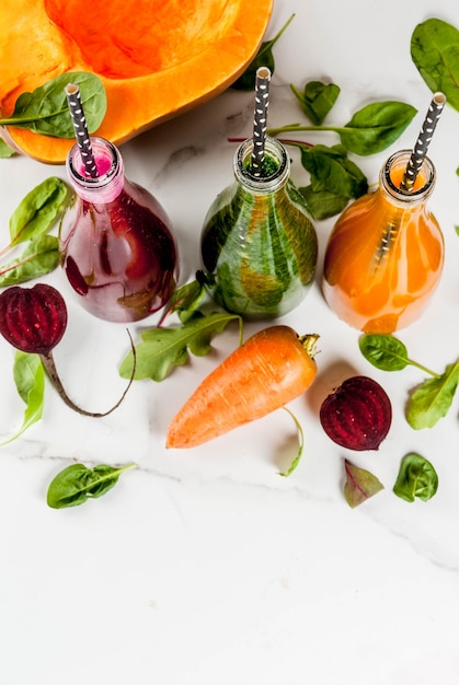 Vegan diet food. Selection colorful fresh organic vegetable smoothies with autumn vegetables: beetroot, pumpkin, carrot, leafy vegetables. In bottles, with straws, white marble table. Copy space