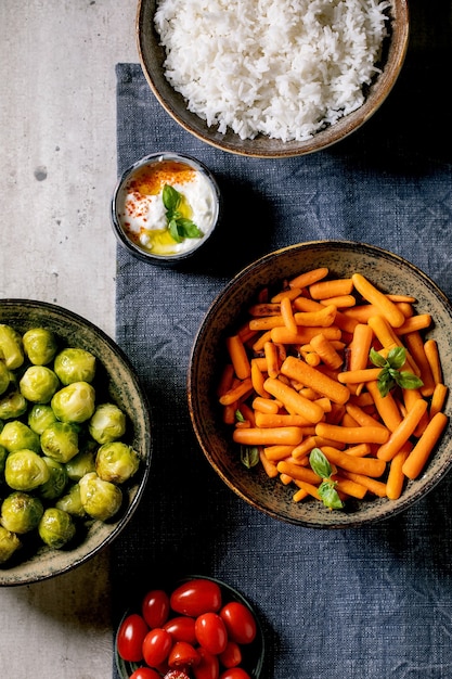 Vegan dinner table with rice and vegetables baked brussel sprouts, baby carrot, cherry tomatoes in ceramic bowls on blue linen tablecloth over grey background. Flat lay.