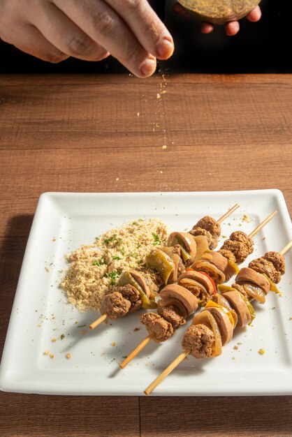 Vegan food cook tossing condiments on vegan skewers on a white\
plate over wood selective focus