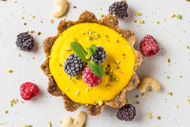 Vegan food dessert. raw vegan yellow cashew mango tart with frozen berries, pistachios and mint. close up