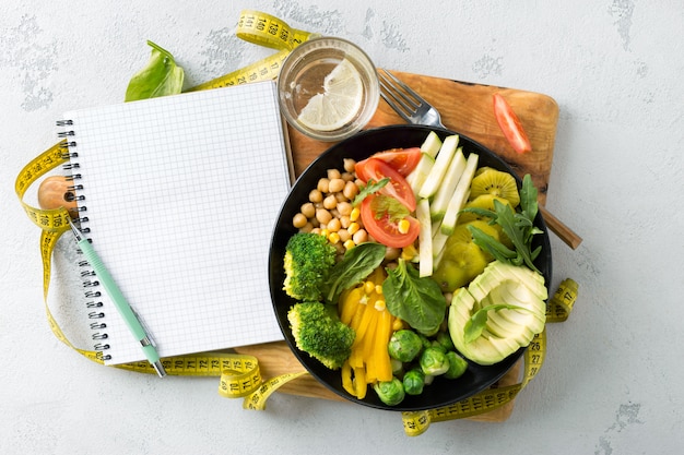 Vegan healthy balanced diet. Vegetarian buddha bowl with blank notebook and measuring tape. ÃÂ¡hickpeas, broccoli, pepper, tomato, spinach, arugula and avocado in plate . Top v