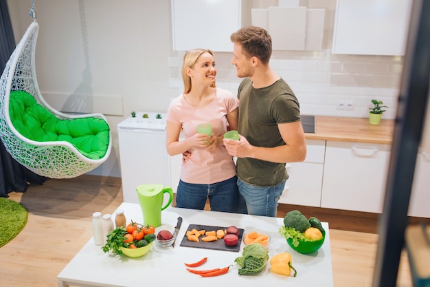 Vegan loving couple holds fresh detox drinks while cooking raw vegetables in the kitchen