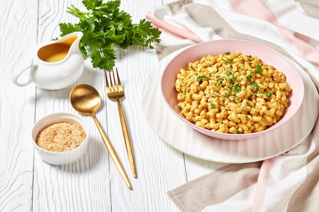 Vegan mac en kaas met voedingsgistsaus in een roze kom op een witte houten tafel met gouden bestek, landschapsmening van bovenaf, close-up