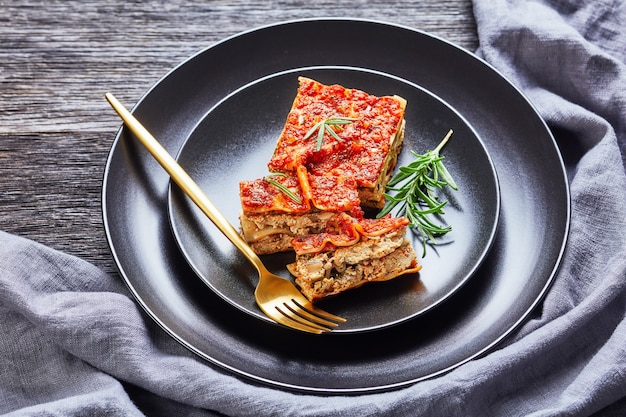 Vegan tofu spinach lasagna with champignon and tomato vegan
bolognese and italian seasoning served on a black plate with fork
and fresh rosemary on a dark rustic wooden table, close-up