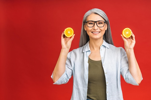 Vegan or vegetarian concept Portrait of a beautiful elderly asian mature aged woman holding an orange fruit smiling isolated over red background