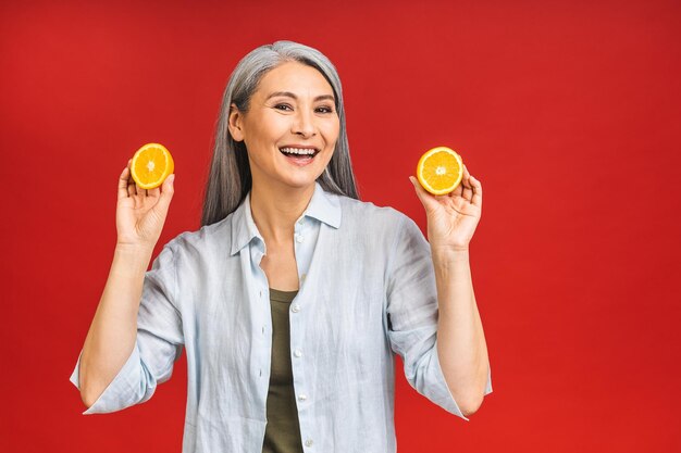 Vegan or vegetarian concept Portrait of a beautiful elderly asian mature aged woman holding an orange fruit smiling isolated over red background