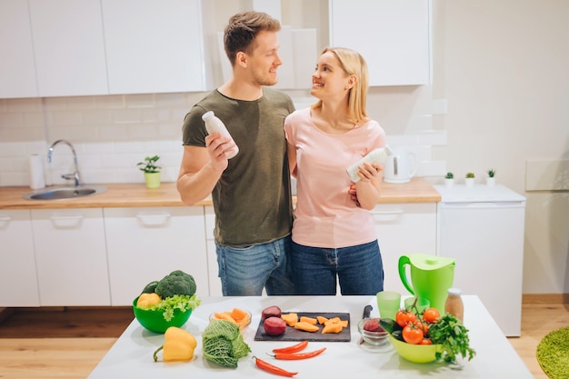 Veganistisch gelukkig liefdevol stel houdt flessen met natuurlijke smoothie vast tijdens het koken van rauwe groenten in de witte keuken. dieet ontgiften. gelukkig gezin. rauw voedsel dieet. vegetarisch eten