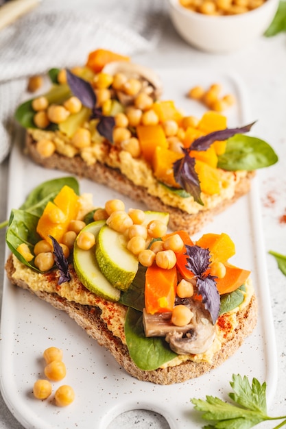 Veganistische gezonde sandwiches met hummus, kikkererwten, gebakken groenten en basilicum op een witte plaat.