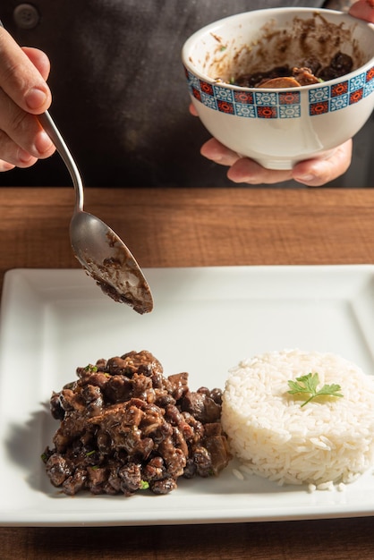Veganistische voedselkok die veganistische feijoada-rijst en farofa op een witte plaat boven hout selectieve focus zet