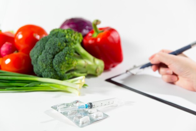 Vegetable diet nutrition or medicaments concept. Female person hands writing diet plan, ripe vegetable composition, drugs and syringe isolated on white background