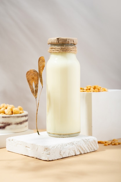 Vegetable pea milk in a bottle and chick peas in handmade ceramic bowl on white podium, pedestal on beige background. shadows. dry leaves. Gluten-free, lactose-free vegan product. Modern. Copy space