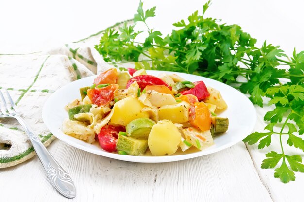 Vegetable ragout with zucchini, cabbage, potatoes, tomatoes and bell peppers in creamy sauce in plate, kitchen towel, parsley and a fork on the background of light wooden board