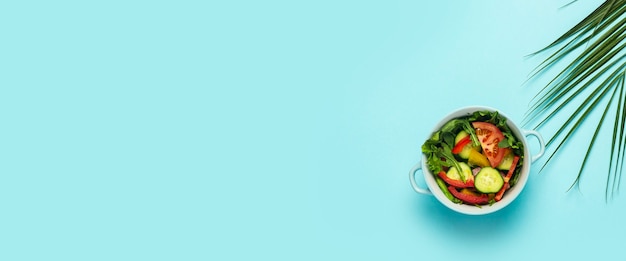 Vegetable salad in a blue bowl and a green leaf of a palm tree on a blue background.