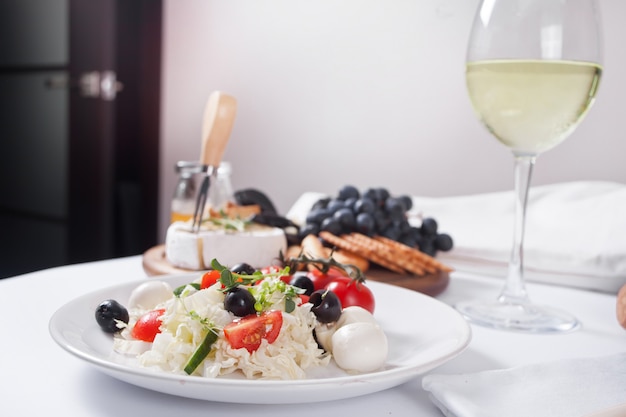 Овощной салат на тарелке, стакан белого вина и тарелка с ассорти из сыра, фруктами и другими закусками.
