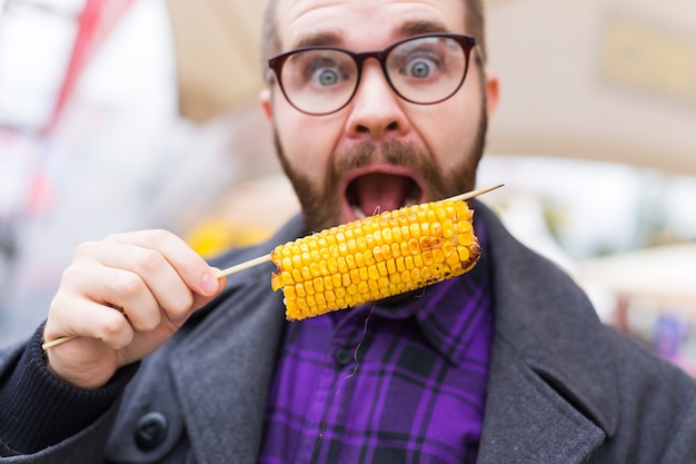 写真 ベジタリアンと食事のコンセプト – ファストフードフェスティバルで屋台のトウモロコシを食べるハンサムな男