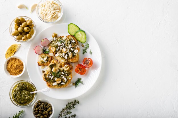 Vegetarian food concept Cauliflower steak with spices chimichurri sauce almond flakes olives fried cherry tomatoes and capers on a white plate White background