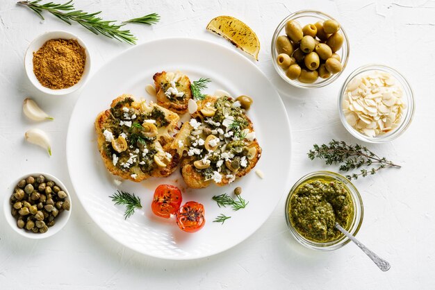 Vegetarian food concept Cauliflower steak with spices chimichurri sauce almond flakes olives fried cherry tomatoes and capers on a white plate White background