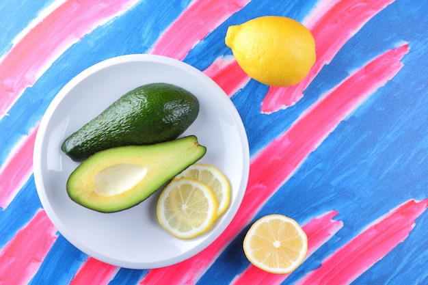 Vegetarian food concept Two halves of a green fresh ripe avocado and a juicy yellow lemon on a white plate on a bluepink background Closeup