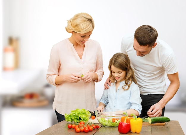 вегетарианская еда, кулинария, счастье и концепция людей - счастливая семья готовит овощной салат на ужин на кухонном фоне