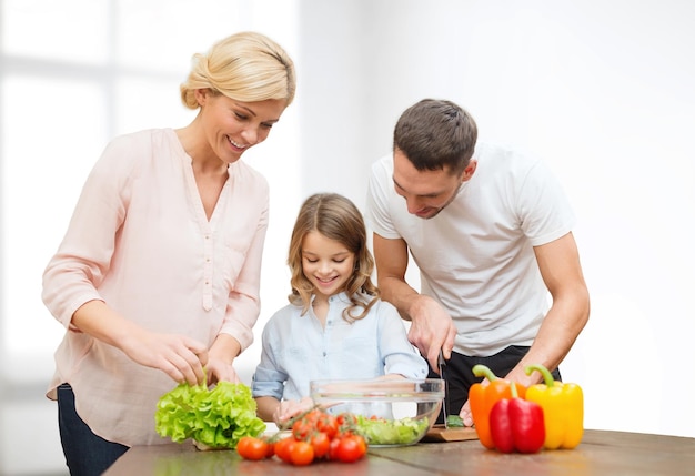 Cibo vegetariano, cucina, felicità e concetto di persone - famiglia felice che cucina insalata di verdure per cena su sfondo bianco della stanza